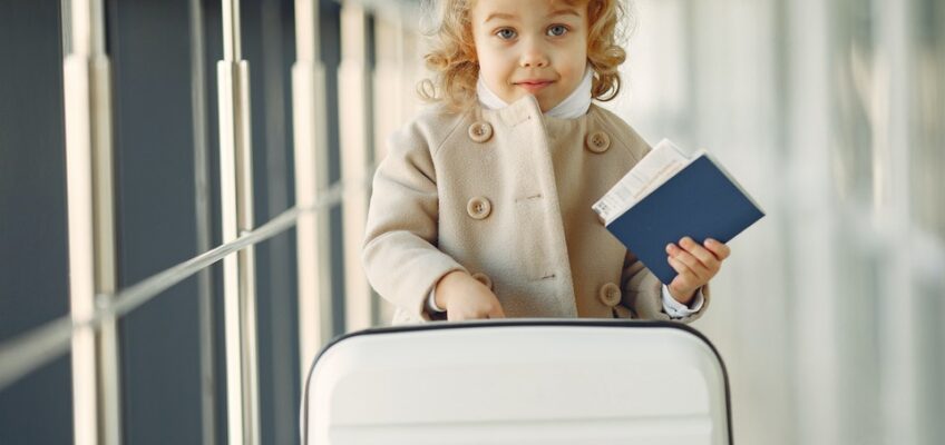 Young child with curly hair holding a passport and standing beside a suitcase in an airport setting.