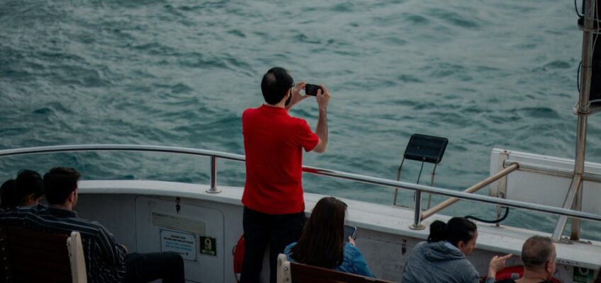 Man in red shirt capturing the sea view with his camera aboard a boat, while other passengers relax and enjoy the voyage.