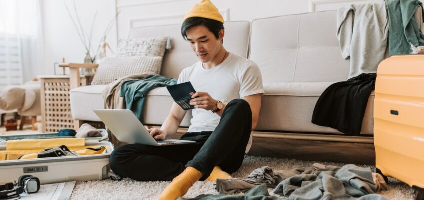 Young man in a yellow beanie sitting on the floor, checking his passport and browsing on his laptop amidst travel preparations, representing research on visa status.