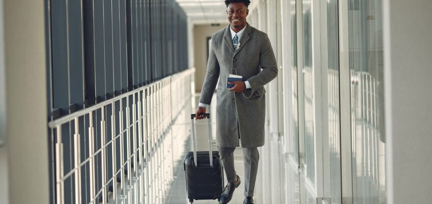 Confident businessman walking in an airport corridor with a suitcase, holding a passport and boarding pass, representing preparation for international travel.