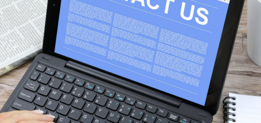 A person using a laptop with a 'CONTACT US' page displayed, alongside a newspaper and a cup of coffee on a wooden table.