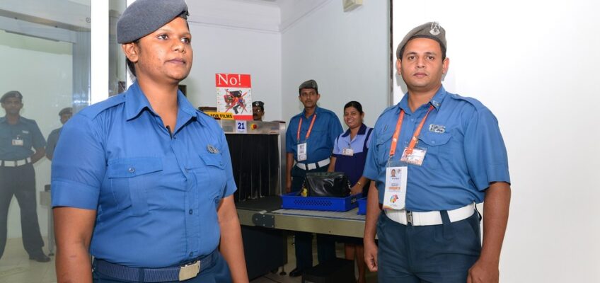 Two security personnel in blue uniforms standing at a checkpoint, with colleagues in the background.