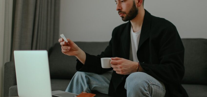 Man in a black jacket holding a credit card and coffee cup, looking at a laptop, ready to apply for an eVisa.