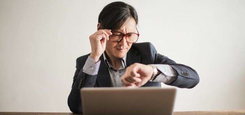 Professional adjusting glasses while intently checking time on his wristwatch, working on a laptop.