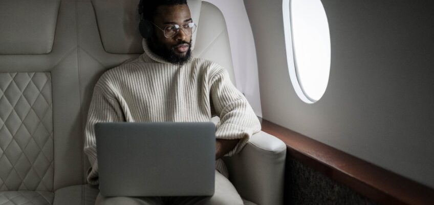 Man in a white sweater working on a laptop while seated in a luxurious private jet interior, looking out through the airplane window.