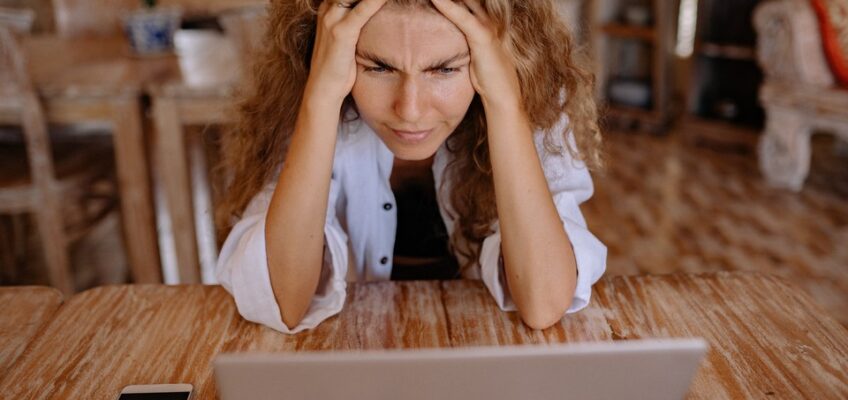 Frustrated woman with curly hair sitting at a wooden table, gripping her head while staring intently at a laptop, symbolizing challenges with online processes.