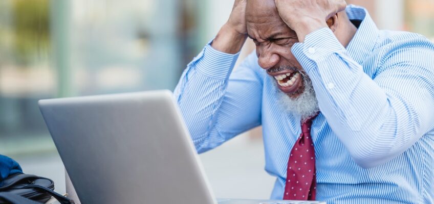 Frustrated man in a blue shirt and red tie holding his head in distress while looking at a laptop, symbolizing visa application challenges.