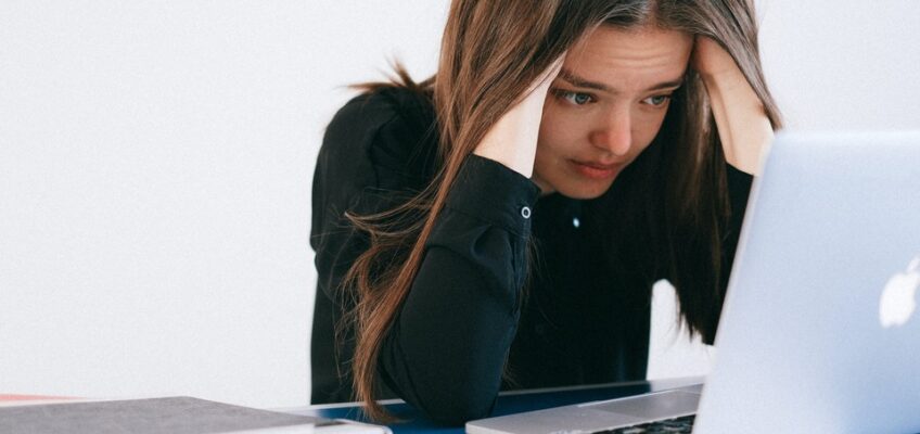 Stressed young woman with long brown hair looking at a laptop screen, holding her head in her hands.