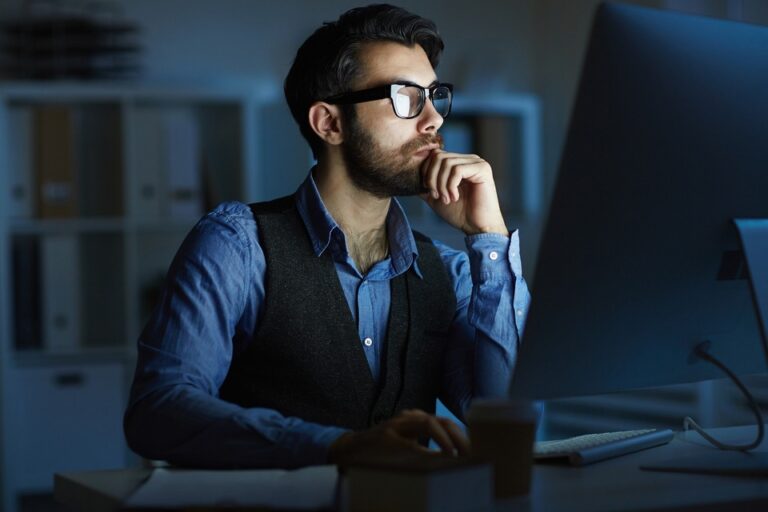 Focused man with glasses working intently at a computer in a dimly lit office.