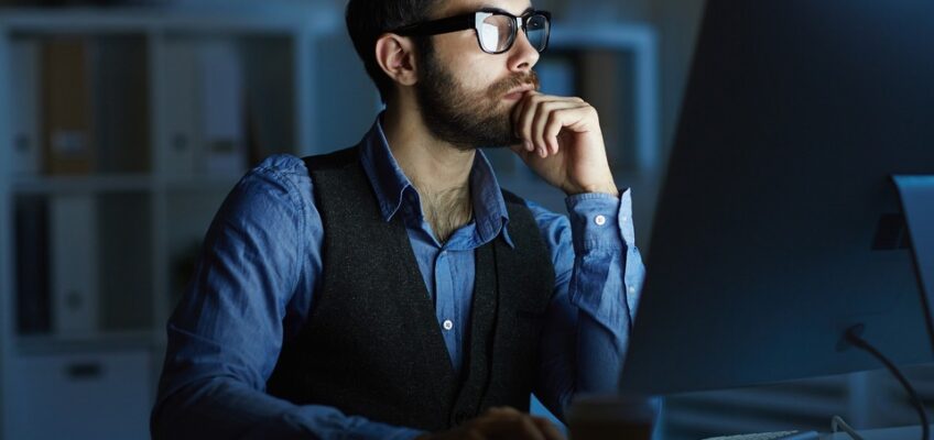 Focused man with glasses working intently at a computer in a dimly lit office.