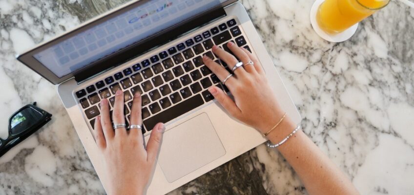 Top view of a person typing on a laptop with a glass of orange juice beside, set on a marble surface.