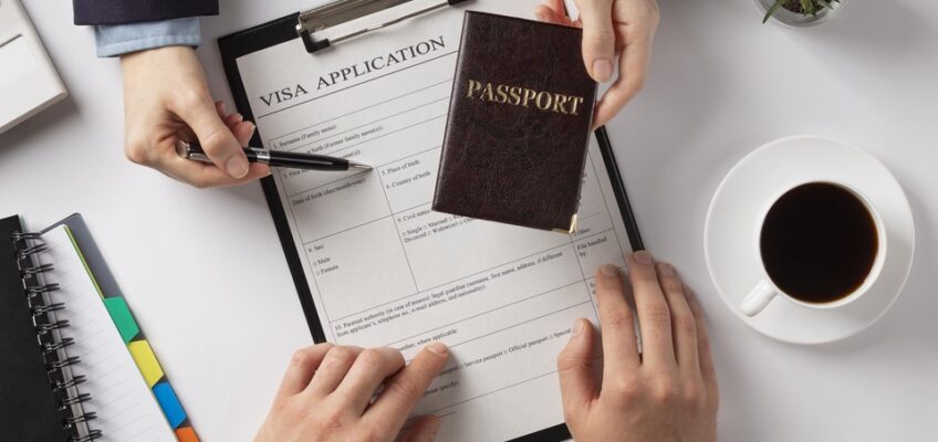 Overhead view of a person's hands holding a pen and a passport over a visa application form, with a notebook, a cup of coffee, and a small potted plant on a white desk.