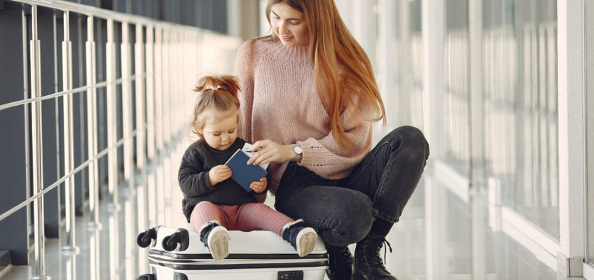 A mother sitting on a suitcase in an airport corridor, showing a passport to her young daughter who is sitting on the suitcase with her.