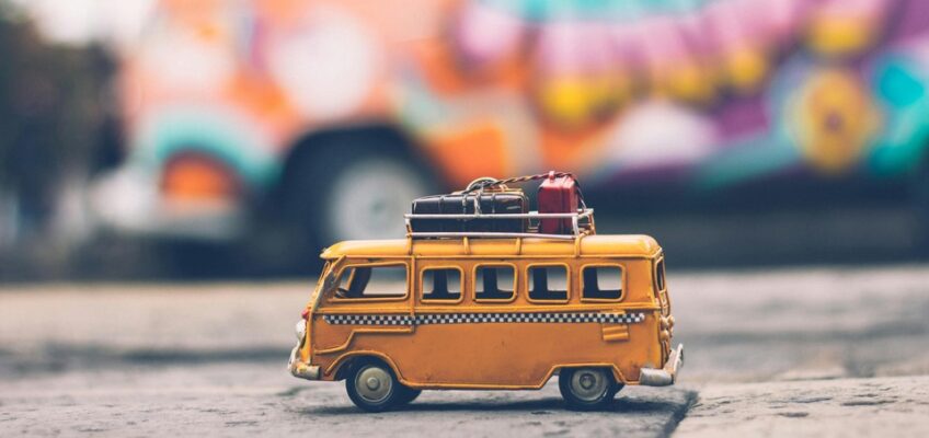 A miniature model of a yellow van with luggage on top is displayed in front of a blurred background with colorful street art.