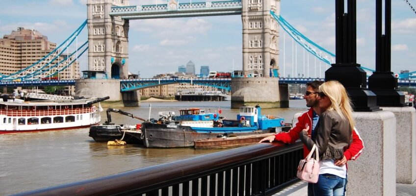 A couple overlooking the iconic Tower Bridge in London, pondering travel regulations, with boats navigating the River Thames below.