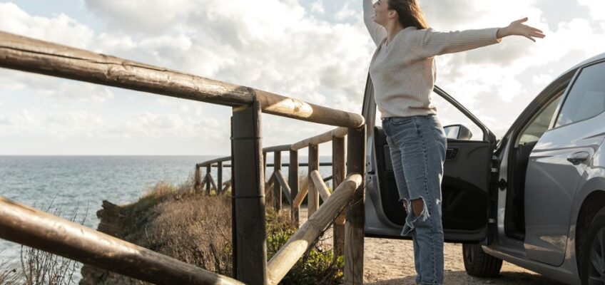 A person standing beside an open car door, arms spread wide, expressing joy and freedom by the seaside, symbolizing the ease of travel with proper authorizations like the New Zealand eTA.