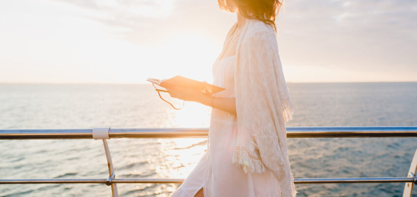 A woman in a white dress and shawl reading on a tablet while leaning on the railing of a cruise ship, symbolizing preparation for sea travel and the importance of electronic travel authorization.