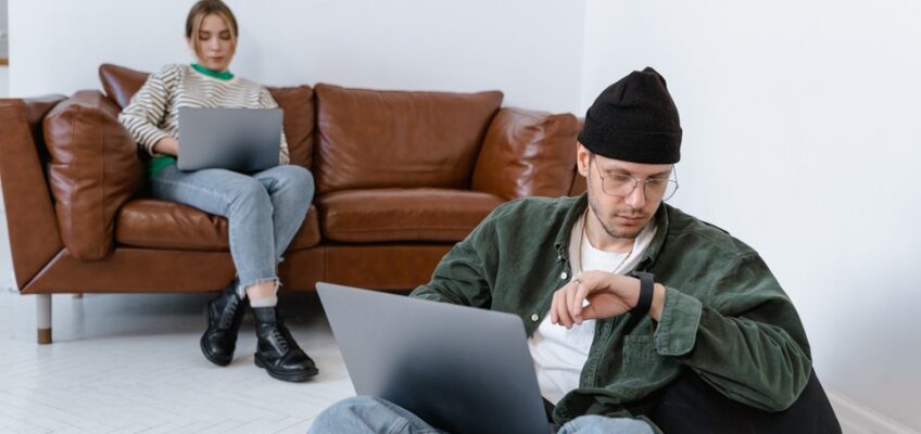 A focused man sitting with a laptop perched on his knees, while a woman sits behind him on a brown leather sofa, engrossed in her own laptop, symbolizing the wait and anticipation in processing applications like ETIAS.