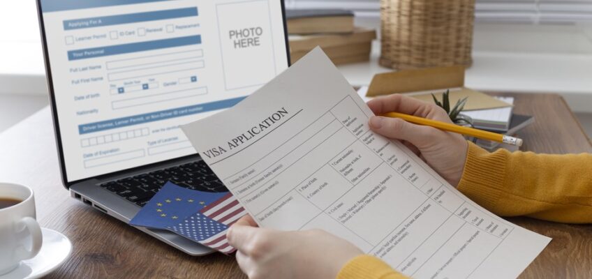 A person filling out a visa application form with a pencil, with a laptop displaying a visa application screen, a cup of coffee, a European Union passport, and a plant in the background.