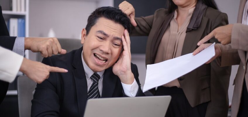 A man appears distressed at his computer while multiple colleagues point at him and his screen, one presenting a document, symbolizing the stress and confusion that might arise from a New Zealand eTA not being approved.