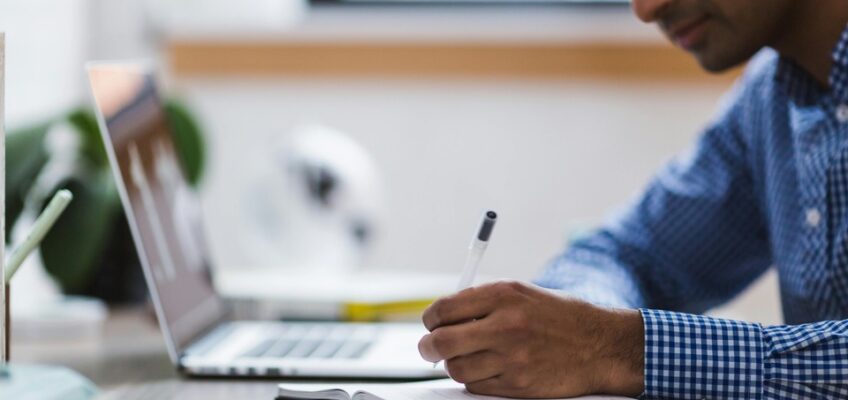 Man in a checkered shirt taking notes in a notebook from information on a laptop, possibly researching or correcting mistakes for an application process.