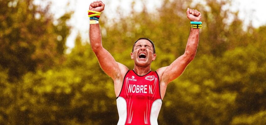 A triumphant triathlete raises his fists in victory, adorned with colorful wristbands, against a blurred backdrop of greenery, exemplifying the joy of permitted sports activities under the ETIAS.