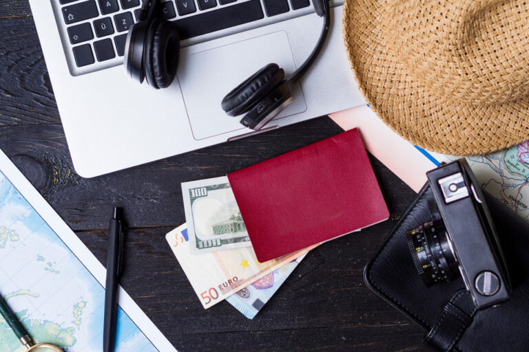 A travel planning workspace with a laptop, headphones, a straw hat, a red passport, euros, a pen, a camera, and a map, symbolizing the essentials for a New Zealand eTA application.