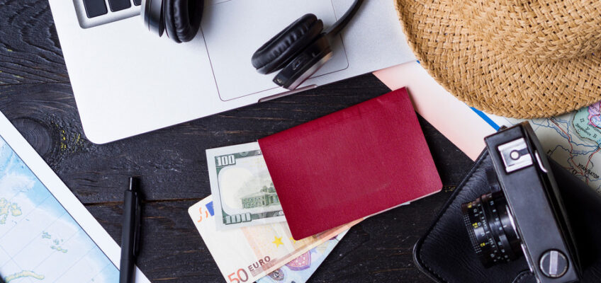 A travel planning workspace with a laptop, headphones, a straw hat, a red passport, euros, a pen, a camera, and a map, symbolizing the essentials for a New Zealand eTA application.