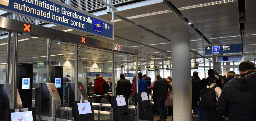 An airport's automated border control area with electronic gates and a sign reading "Automatische Grenzkontrolle / automated border control 18+," with travelers queuing for entry.