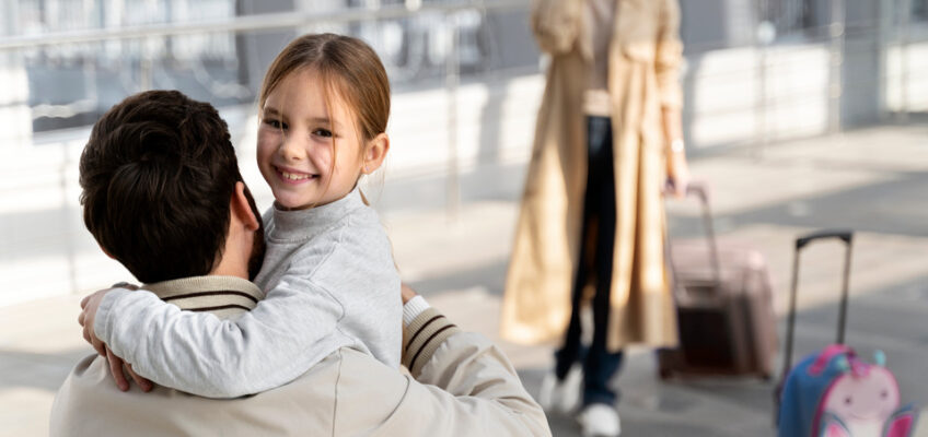 A happy child hugging her father at the airport, with her mother on a phone call in the background, and a suitcase and a pink butterfly-themed backpack indicating travel.