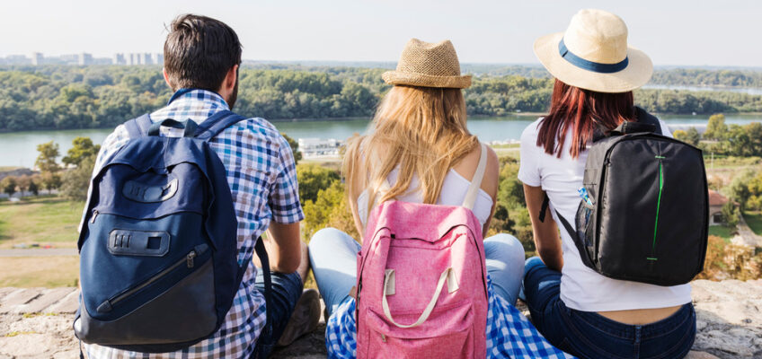 Three travelers sitting side by side overlooking a river, each with a backpack, indicating companionship and travel.