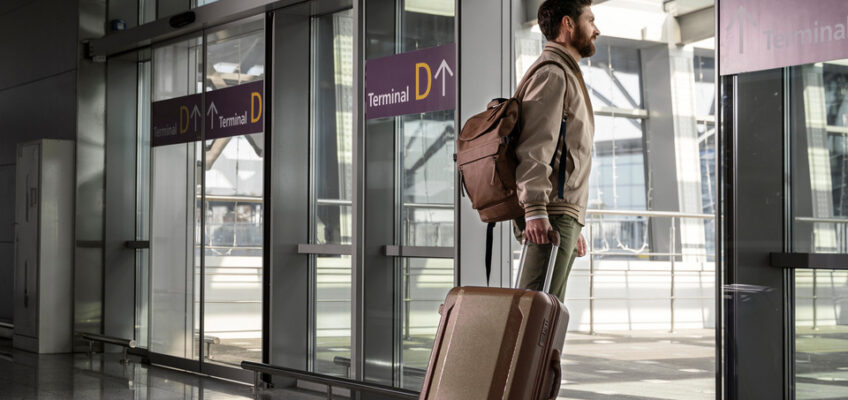 A traveler at an airport terminal, with a suitcase and a backpack, looks out through the glass doors, reflecting the contemplation of journeying with the convenience of a Turkish eVisa.