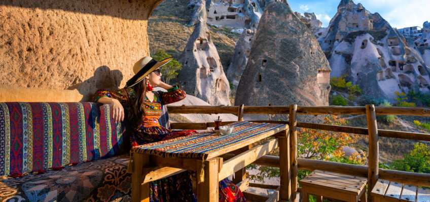 A traveler in traditional attire relaxes on a patterned bench, enjoying the view of Cappadocia's unique fairy chimneys and cave dwellings under a clear blue sky.