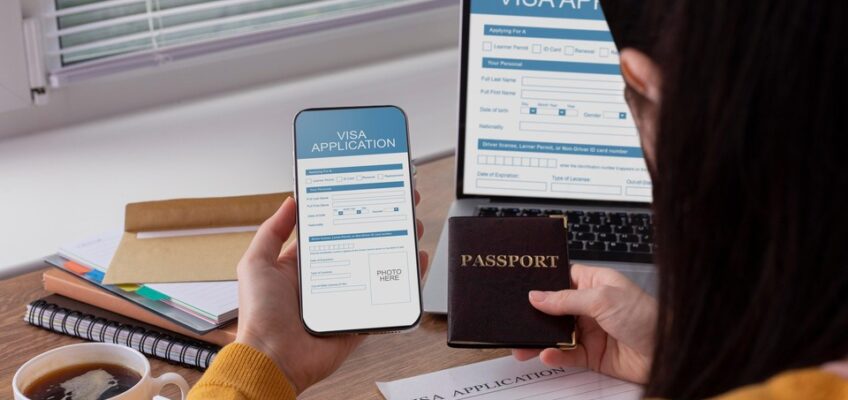 A person holding a passport and a smartphone with a visa application screen open, sitting at a desk with a laptop and a cup of tea, indicating the process of applying for a visa online.