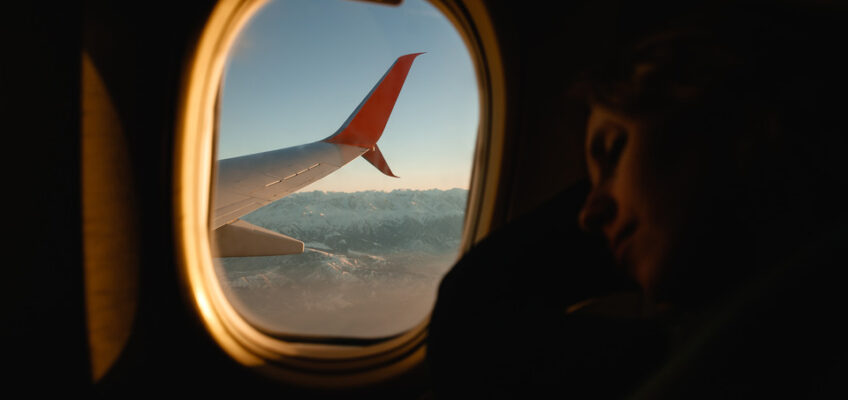 A passenger resting against an airplane window with a view of an airplane wing and mountainous landscape during a flight, reflecting the serene travel experience to New Zealand.