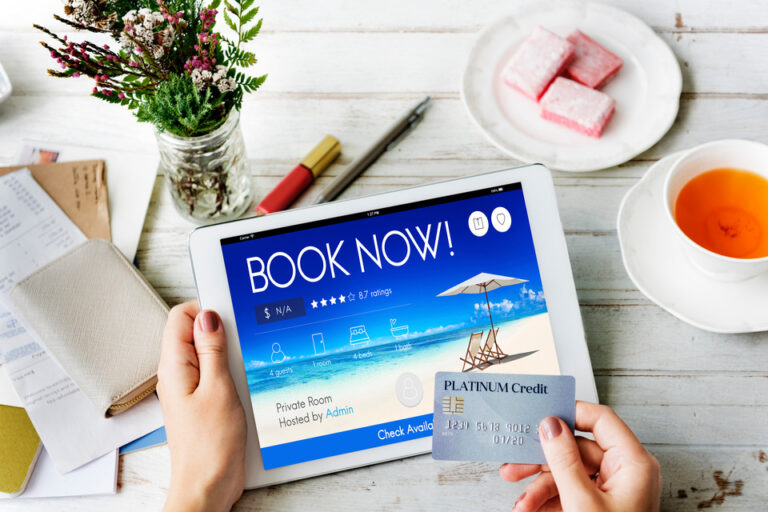 A person's hands holding a credit card and a tablet displaying a "BOOK NOW" screen for vacation booking, with a cup of tea, sweets, and flowers on a wooden table.