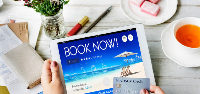 A person's hands holding a credit card and a tablet displaying a "BOOK NOW" screen for vacation booking, with a cup of tea, sweets, and flowers on a wooden table.