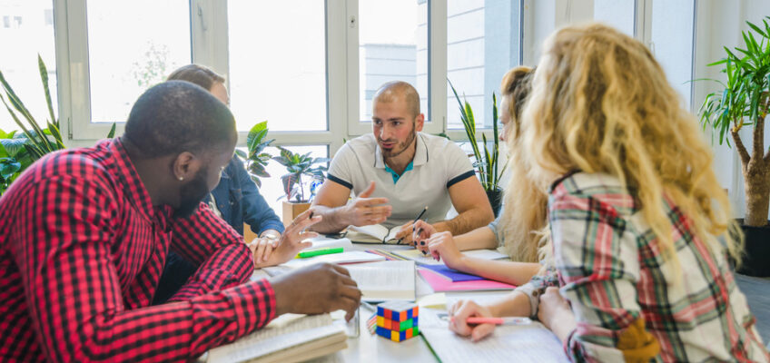 A diverse group of professionals engaged in a collaborative meeting around a table with papers and a colorful puzzle cube, in a bright office space.