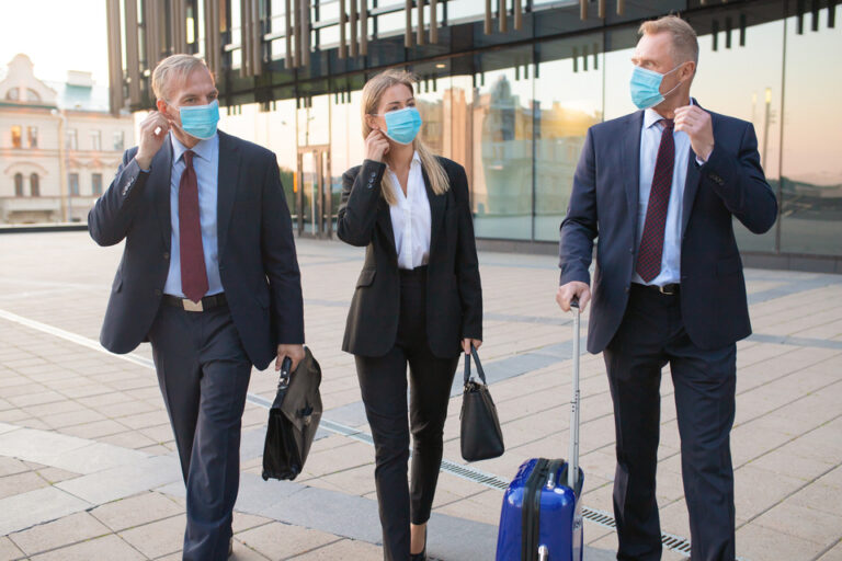 Three business professionals wearing face masks walking outside an airport, reflecting the new norms of travel with health precautions.