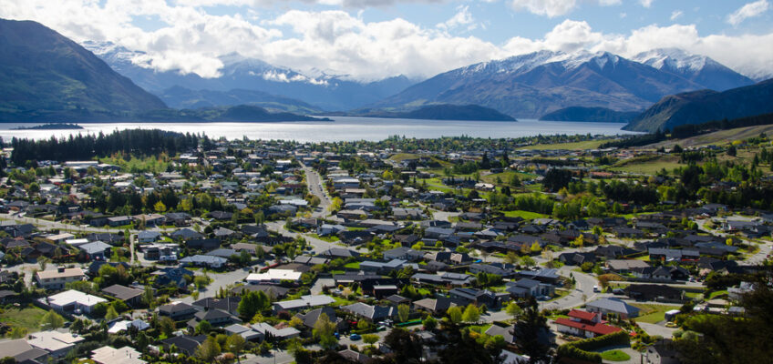 A picturesque view of a New Zealand town with houses spread across a landscape, set against a backdrop of a serene lake and towering mountains.