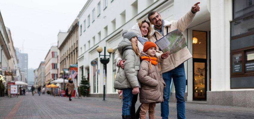 A cheerful family of four, bundled up in warm winter clothes, is exploring a city street together with a map, as the father points out a direction, exemplifying group travel.