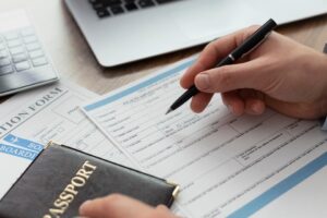 A person completing an application form with a black pen, with a dark-colored passport and a laptop in the background.