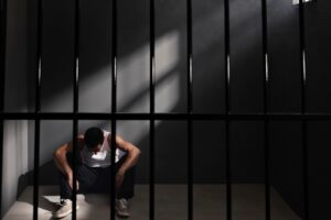 A despondent man sitting on the floor behind prison bars, reflecting on the challenges of applying for travel authorization with a criminal history.