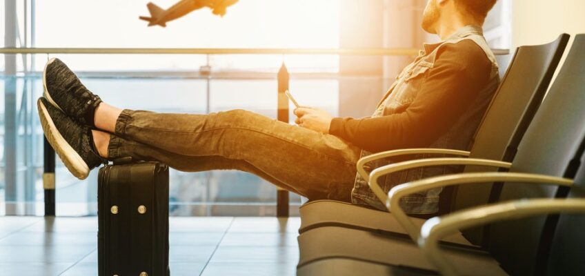 A relaxed traveler sits with legs on a suitcase, using a smartphone in the airport lounge with a view of an airplane taking off in the background.