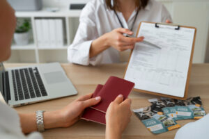 A person holding a red passport across a desk from a travel consultant, who is pointing to a form on a clipboard, with travel pamphlets and a laptop on the desk, illustrating the process of applying for a U.S. ESTA.