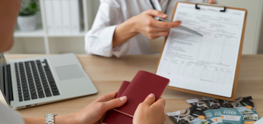 A person holding a red passport across a desk from a travel consultant, who is pointing to a form on a clipboard, with travel pamphlets and a laptop on the desk, illustrating the process of applying for a U.S. ESTA.