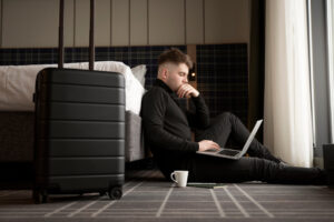 A focused man sitting on the floor of a hotel room, with a laptop on his legs and a suitcase nearby, as he appears to be checking his travel documents online.