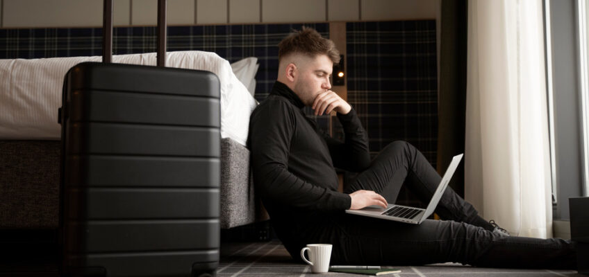 A focused man sitting on the floor of a hotel room, with a laptop on his legs and a suitcase nearby, as he appears to be checking his travel documents online.