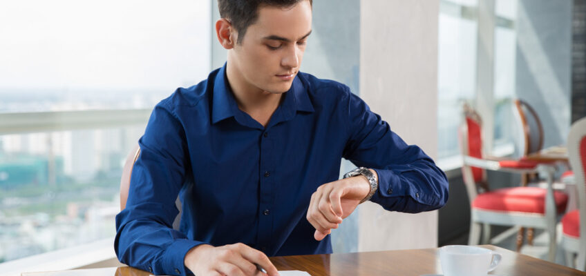 A man in a blue shirt checks his watch while sitting at a cafe table, symbolizing the concern for timely eVisa application processing.