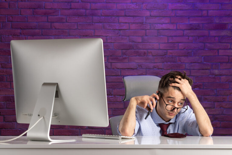 Stressed businessman with glasses looking exasperated while on the phone, sitting in front of a computer screen, symbolizing frustration with the U.S. ESTA website issues.